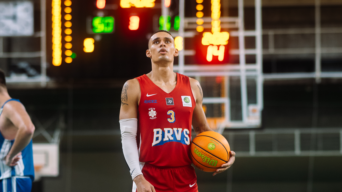 Joueur de basketball en tenue rouge BRVS n°3 tenant un ballon, debout devant un panneau d’affichage lumineux en salle.