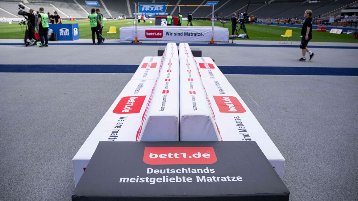 Photo prise depuis le bord du terrain dans un stade, au-dessus d'un banc de touche portant le logo bett1 sur des stations d'athlétisme au milieu du terrain.