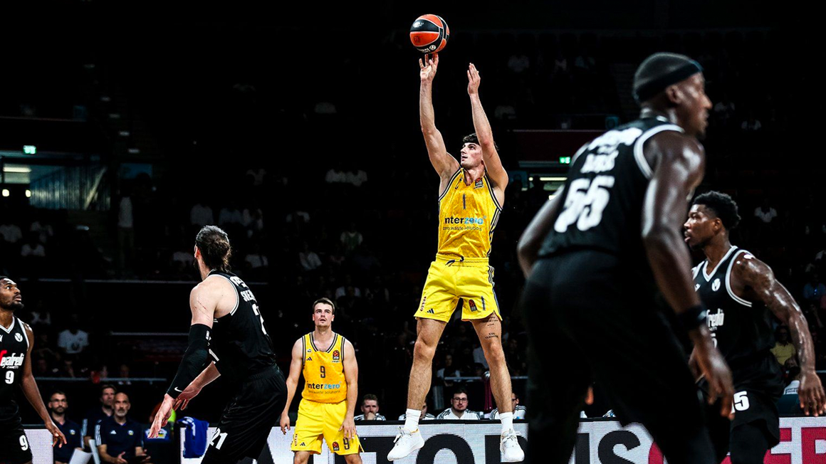 Photo : Un terrain de basket où deux équipes s'affrontent. Un joueur en plein saut lance le ballon pour marquer