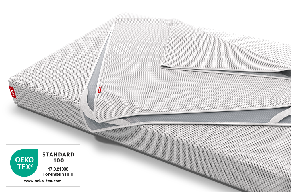Der BODYGUARD® Nässeschutz für Kindermatratzen liegt zusammengelegt auf der bett1 Babymatratze, daneben das Oeko-Tex® Standard 100 Siegel.