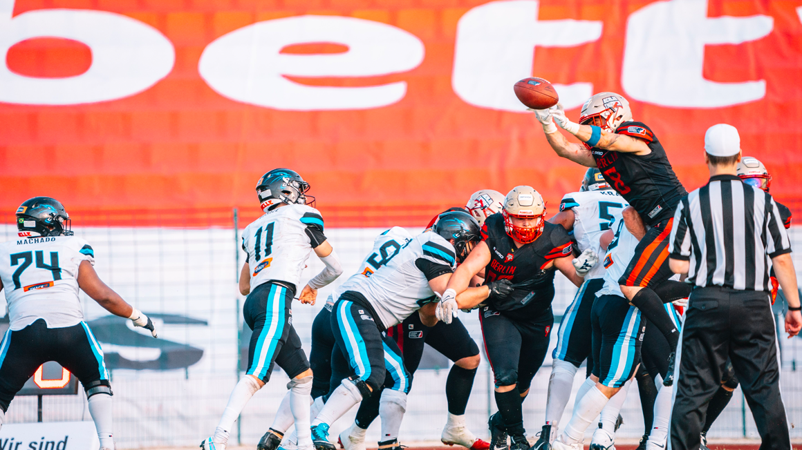 Deux équipes de football américain et un joueur saute pour rattraper le ballon. En fond, le logo bett1 en affiche