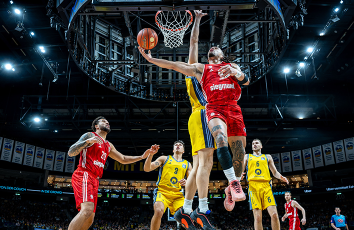 Foto: Am Spielfeldrand stehen die Spieler von ALBA Berlin um eine sitzende Person im Anzug herum.
