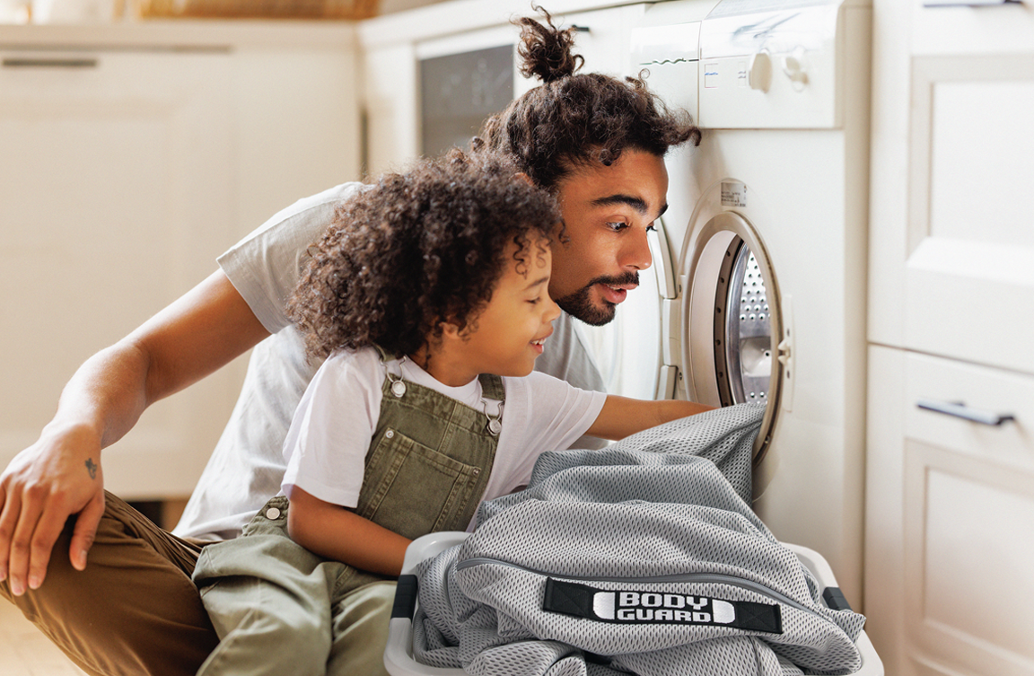 Un père et sa fille regardent à l’intérieur d’un lave-linge, avec une housse BODYGUARD® posée devant.
