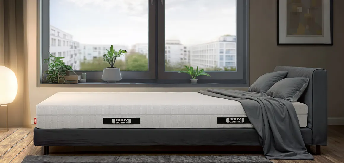 Matelas BODYGUARD® sur un lit gris dans une chambre moderne avec grande fenêtre et vue sur la ville.