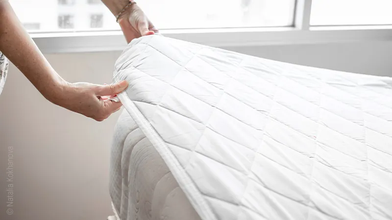 Foto: Zwei Hände greifen einen Matratzenschoner, der auf einer Matratze liegt an einer Ecke