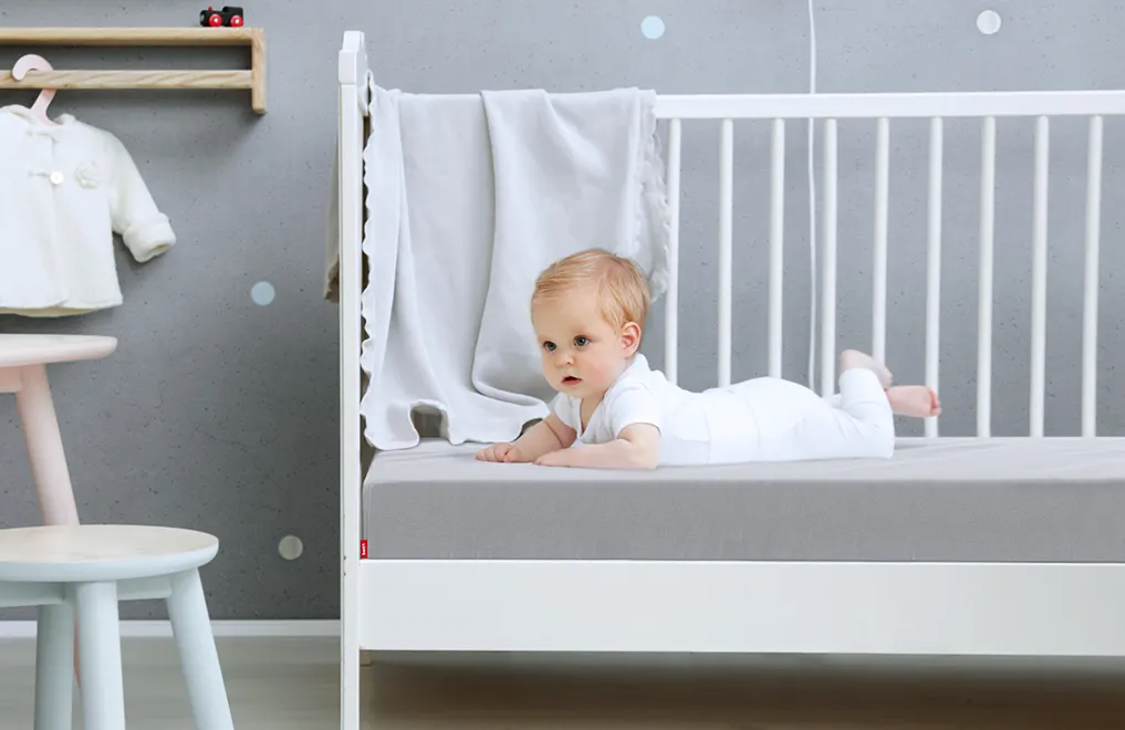 Un bébé est allongé sur le ventre la tête redressée sur un lit à barreau. Le matelas BODYGUARD enfant est recouvert du drap-housse gris enfant.