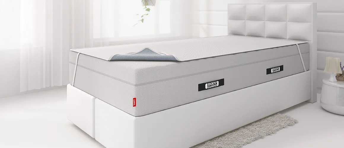 Matelas BODYGUARD® dans une chambre lumineuse, avec surmatelas partiellement replié et étiquette bett1 visible.
