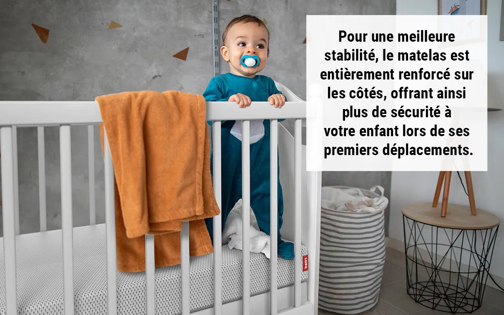 Bébé debout dans un lit à barreaux, tenant le bord. Un matelas BODYGUARD® est visible avec une housse renforcée. Un encart indique que les côtés du matelas assurent plus de sécurité lors des premiers déplacements.