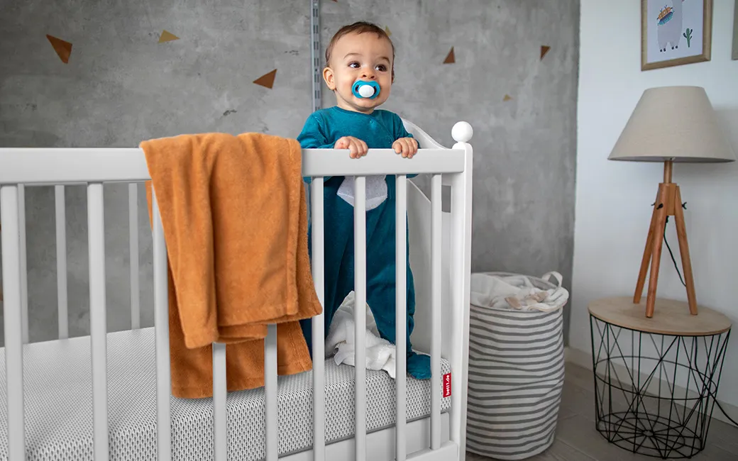 Bébé debout dans un lit à barreaux, tenant le bord. Un matelas BODYGUARD® est visible avec une housse renforcée. Un encart indique que les côtés du matelas assurent plus de sécurité lors des premiers déplacements.