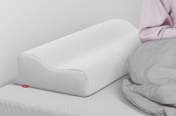 Un oreiller ergonomique bodyguard à texture alvéolée est posé sur un lit, avec sa forme ondulée clairement visible. À droite, une personne assise, partiellement couverte par une couette grise, apparaît hors champ.