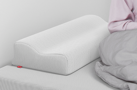 Un oreiller ergonomique bodyguard à texture alvéolée est posé sur un lit, avec sa forme ondulée clairement visible. À droite, une personne assise, partiellement couverte par une couette grise, apparaît hors champ.
