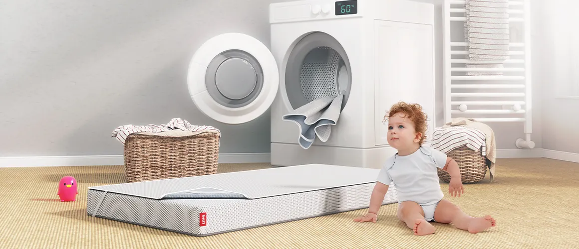 Bébé assis devant un matelas BODYGUARD® ouvert en plusieurs couches, près d’un lave-linge affichant 60 °C. Un encart indique que la housse et la surface de couchage sont lavables à 60 °C.