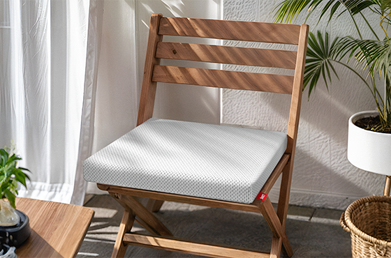 Coussin de chaise BODYGUARD® blanc à housse respirante, posé sur une chaise pliante en bois. L’ensemble est installé dans un espace lumineux avec des plantes et un sol en carrelage. Étiquette rouge bett1 visible.