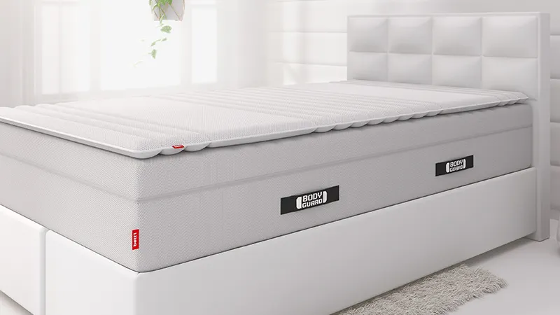 Surmatelas BODYGUARD® posé sur un matelas BODYGUARD® dans une chambre claire au style épuré.