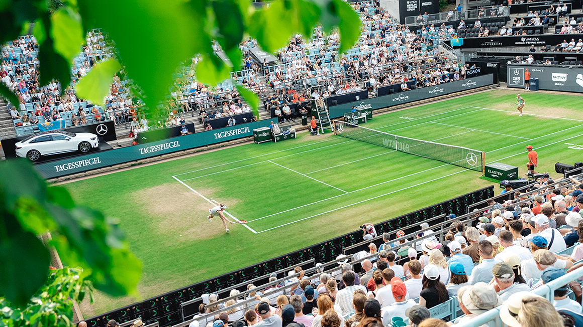 Blick von der Tribüne auf ein Tennisfeld, auf dem ein Tennisspiel stattfindet. Die Tribünen sind voll besetzt.