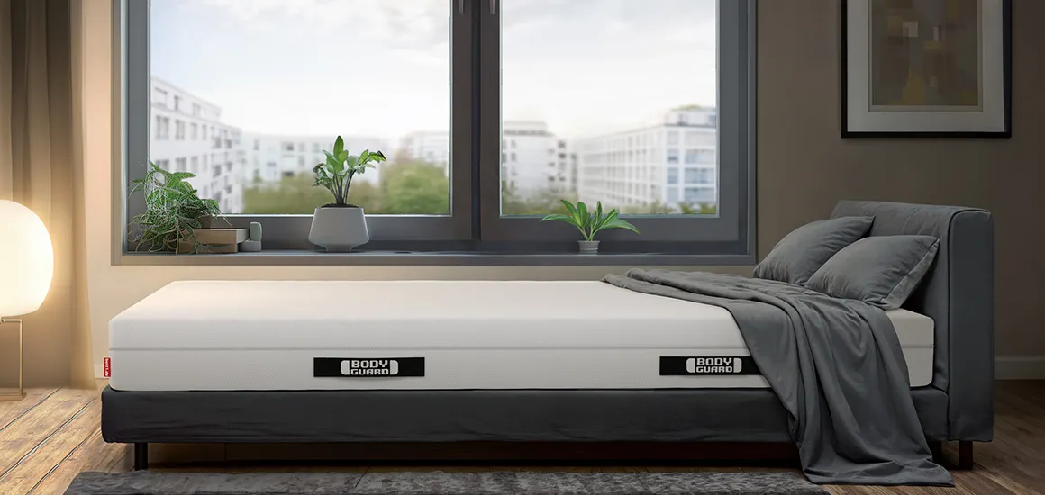 Matelas BODYGUARD® sur un lit gris dans une chambre moderne avec grande fenêtre et vue sur la ville.