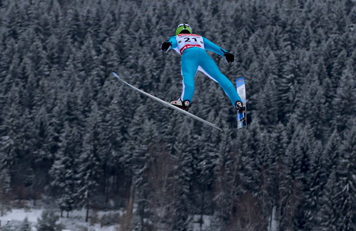 Skieur en combinaison bleue effectuant un saut à ski, vu de dos en plein vol au-dessus d’une forêt enneigée.