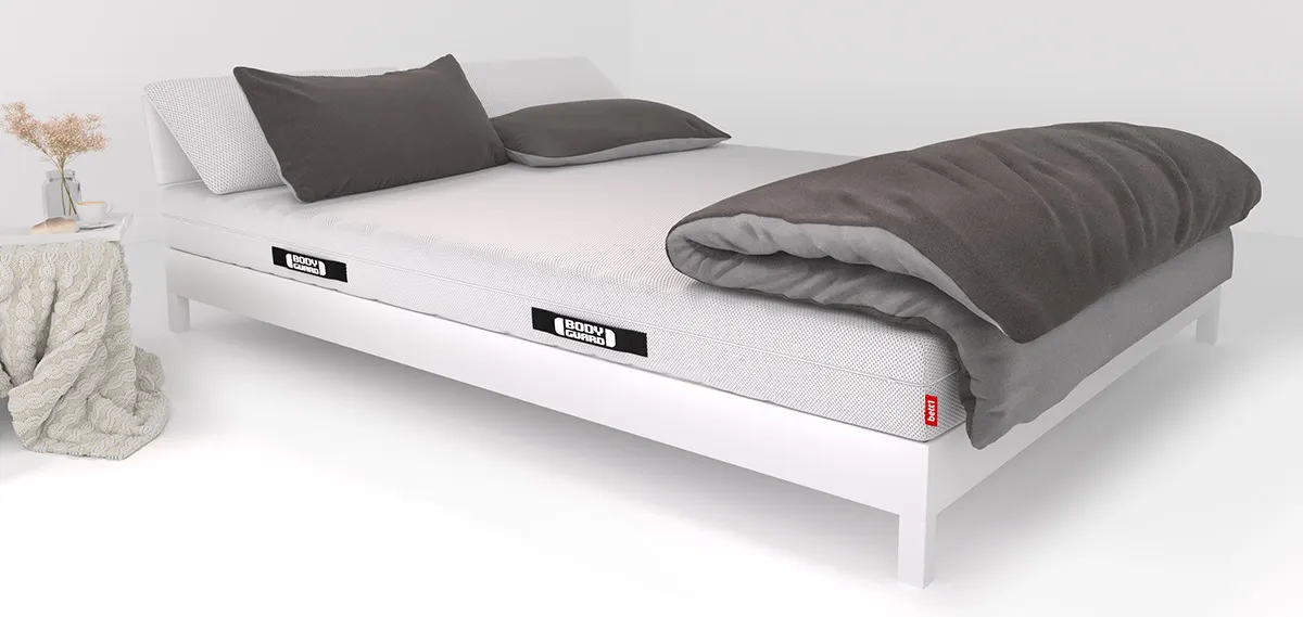 Matelas BODYGUARD® posé sur un cadre de lit blanc avec draps gris foncé, deux oreillers et une couette. Le logo BODYGUARD est visible sur le côté du matelas. Un plaid beige est plié sur une table d’appoint.