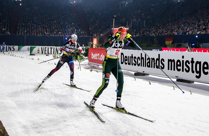 Compétition de biathlon avec deux athlètes en pleine course sur neige, devant une foule en tribunes. Les panneaux publicitaires affichent notamment le logo bett1.de et la mention « Deutschlands meistgekaufte Matratze ».