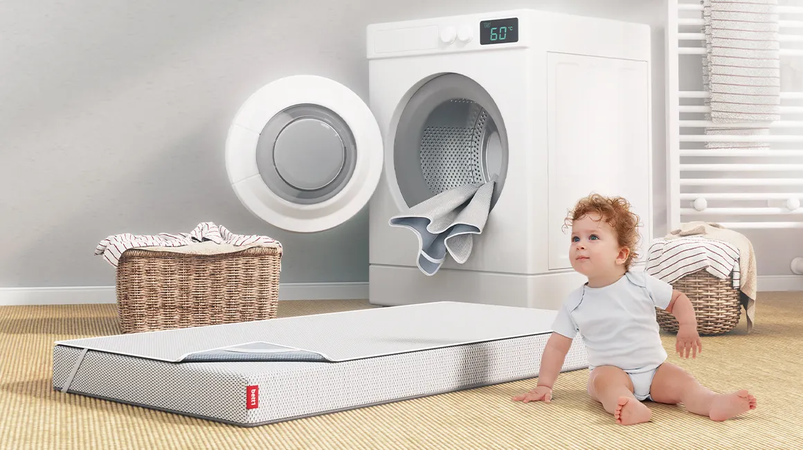 In einem Badezimmer sitzt ein Kleinkind vor einer Waschmaschine. Daneben liegt die SuperBreeze Kindermatratze auf dem Boden. Sie ist mit dem BODYGUARD Nässeschutz bezogen