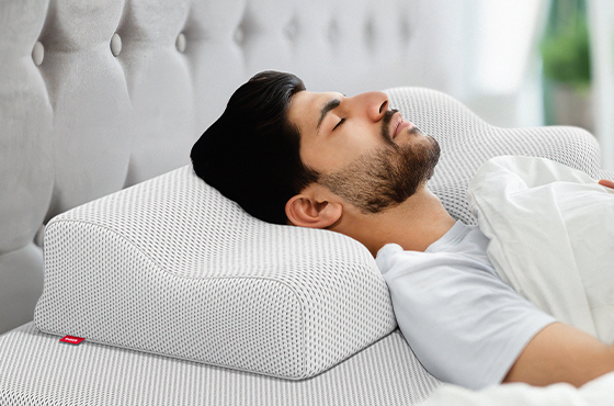 Un homme allongé sur le dos dort sur un oreiller ergonomique blanc à structure alvéolée. Sa tête et son cou sont bien soutenus. Le lit et la tête de lit capitonnée apparaissent en arrière-plan.