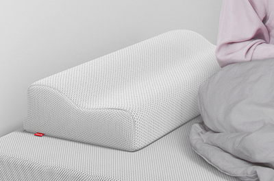 Un oreiller ergonomique bodyguard à texture alvéolée est posé sur un lit, avec sa forme ondulée clairement visible. À droite, une personne assise, partiellement couverte par une couette grise, apparaît hors champ.