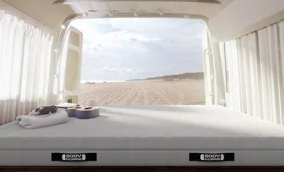 Innenansicht eines Camping-Vans, in dem eine BODYGUARD Matratze 140x190 liegt. Auf der Matratze liegen unter anderem Strandutensilien, Kopfhörer und eine Gitarre. Durch die geöffneten Hintertüren ist ein Strand zu sehen.