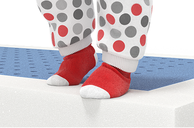 Pieds d’un enfant debout sur un matelas ouvert, portant des chaussettes rouges à bout blanc et un pyjama blanc à pois gris et rouges. Les pieds sont posés sur une surface en mousse bleue perforée.